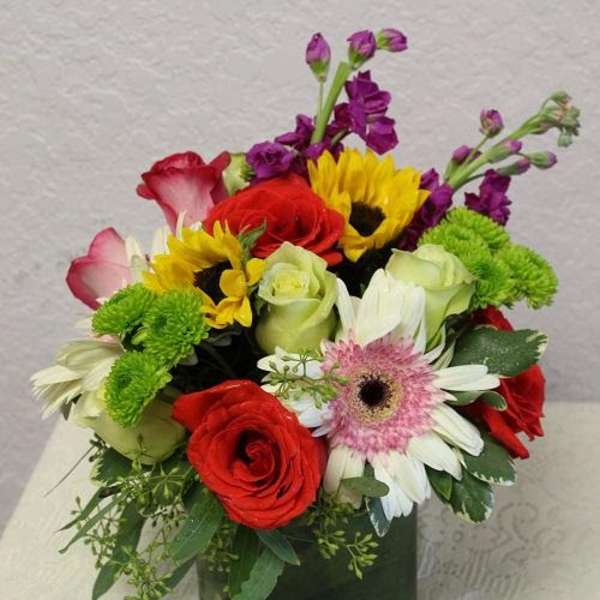 Mixed bouquet of red, yellow, white, and purple flowers in a square glass vase