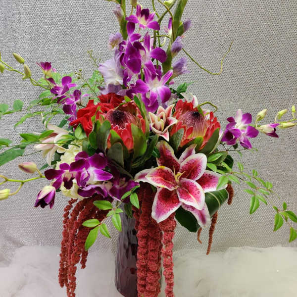 Tall tropical arrangement with purple orchids and pink lilies in a dark vase