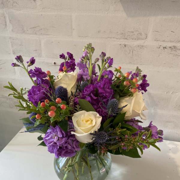Round glass vase of white roses, purple flowers, blue thistles, and orange berries.