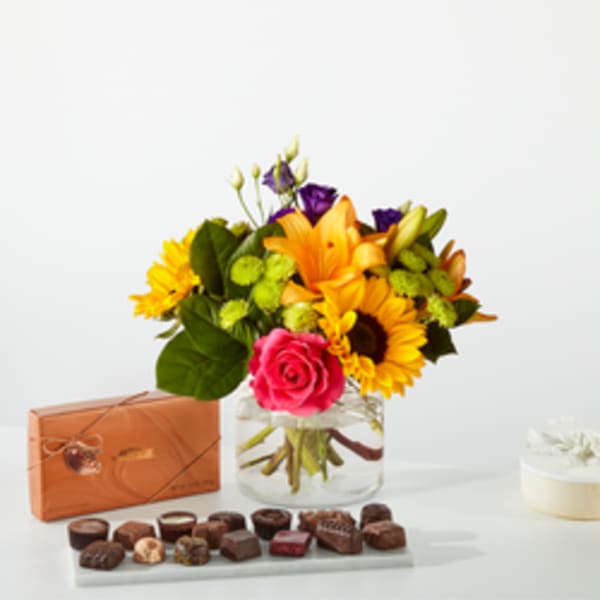 Mixed bouquet in a glass vase with a box of chocolates and a small cake.