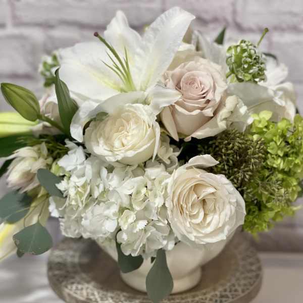 Low white bowl arrangement with white lilies, cream roses, hydrangeas, and green accents