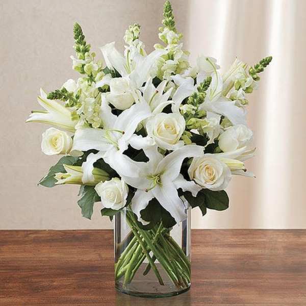 Arrangement of white lilies, roses, and snapdragons in a clear glass cylinder vase on a wooden table.