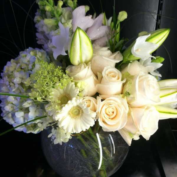Low round arrangement of white roses, lilies, hydrangeas, gerbera daisies, and snapdragons in a clear glass bowl vase