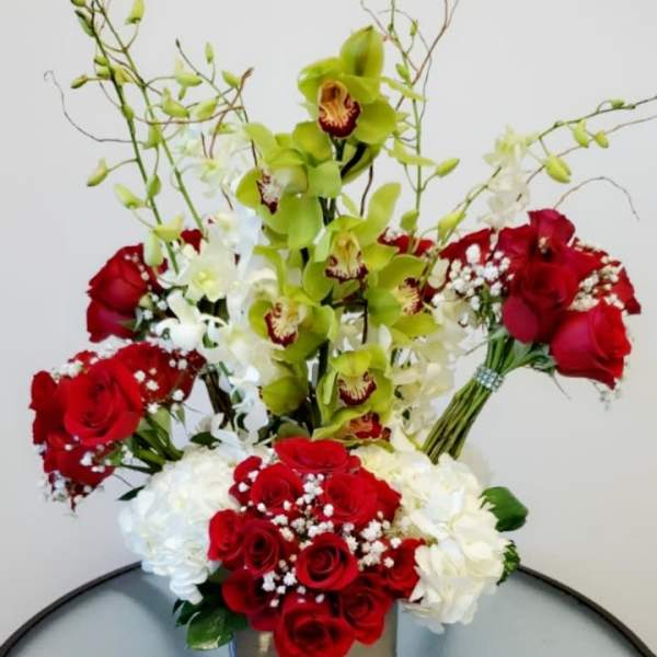 Tall arrangement of red roses, white blooms, and green orchids in a square vase