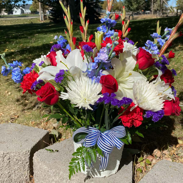 Bouquet of red roses, white lilies, and blue flowers in a white vase with a striped ribbon.