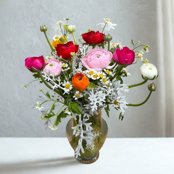Tall mixed bouquet of pink, red, white, and orange ranunculus with small daisies in a green glass vase