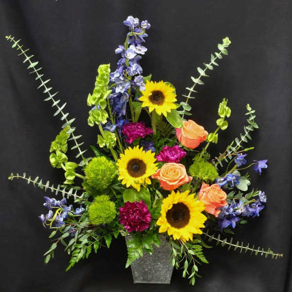 Tall mixed arrangement with sunflowers, orange roses, purple blooms, and blue flowers in a metal vase.