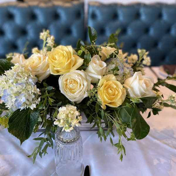 Low arrangement of yellow and cream roses with white hydrangeas on a set table.