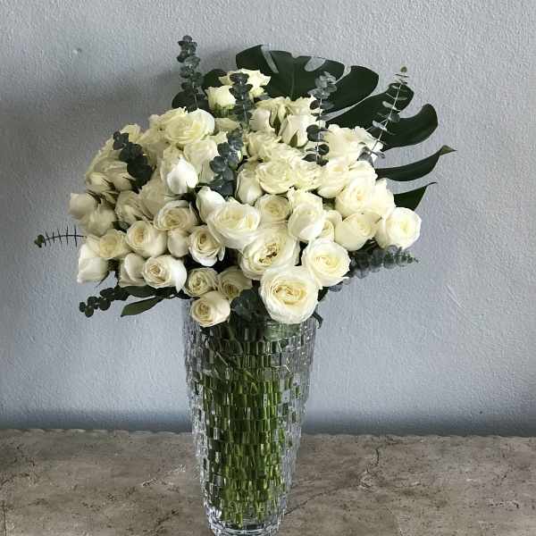 White rose bouquet in a tall clear glass vase with dark green foliage