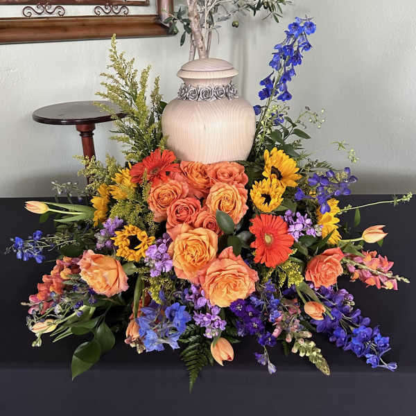 Colorful floral arrangement with roses, gerbera daisies, and blue delphinium around an urn
