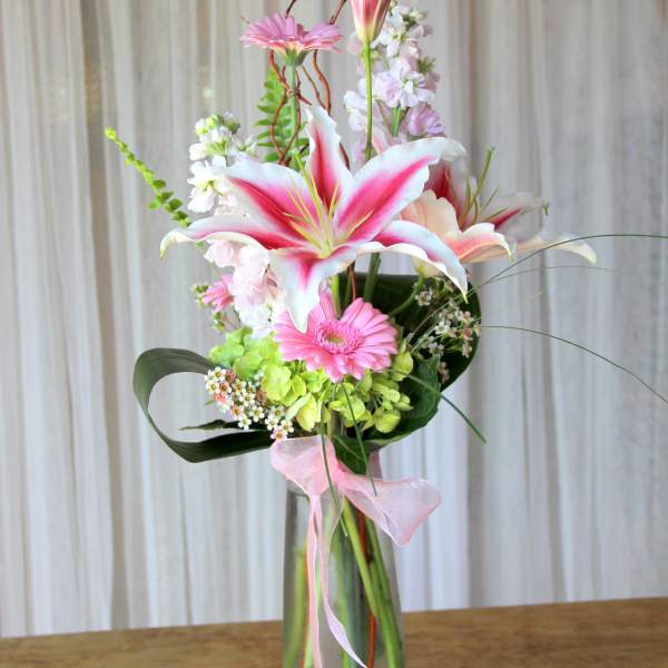 Pink lilies and gerbera daisies in a tall glass vase with a pink ribbon