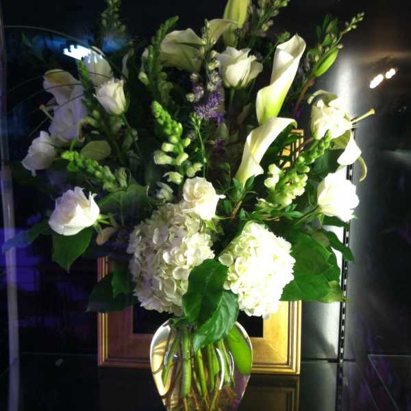 Tall arrangement of white roses, calla lilies, and hydrangeas in a clear glass vase with touches of purple.