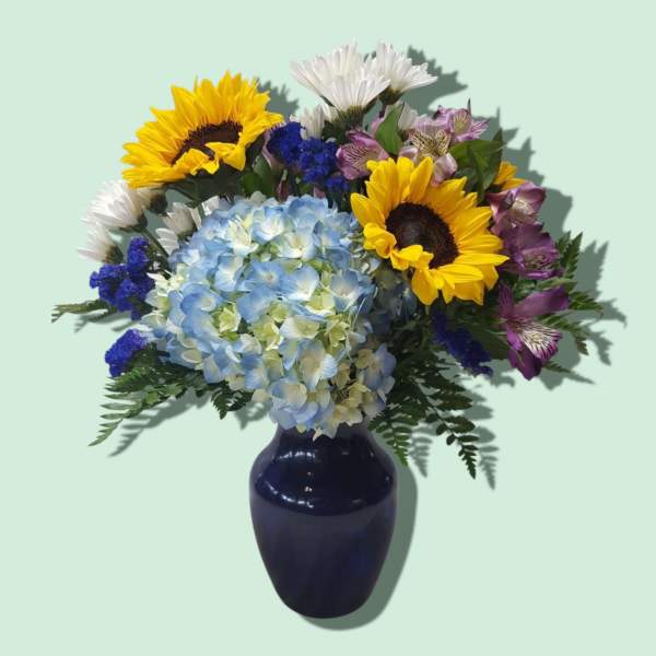 Mixed bouquet with blue hydrangea, yellow sunflowers, white daisies, and purple blooms in a dark blue vase.