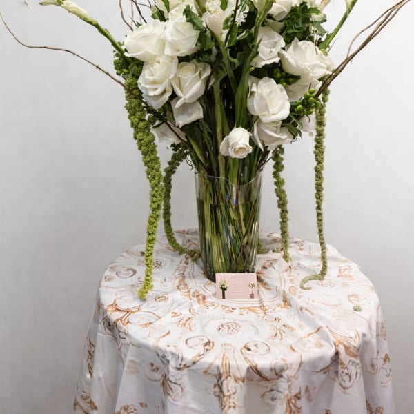 Tall white flower arrangement with roses and trailing green accents in a clear glass vase on a round table.