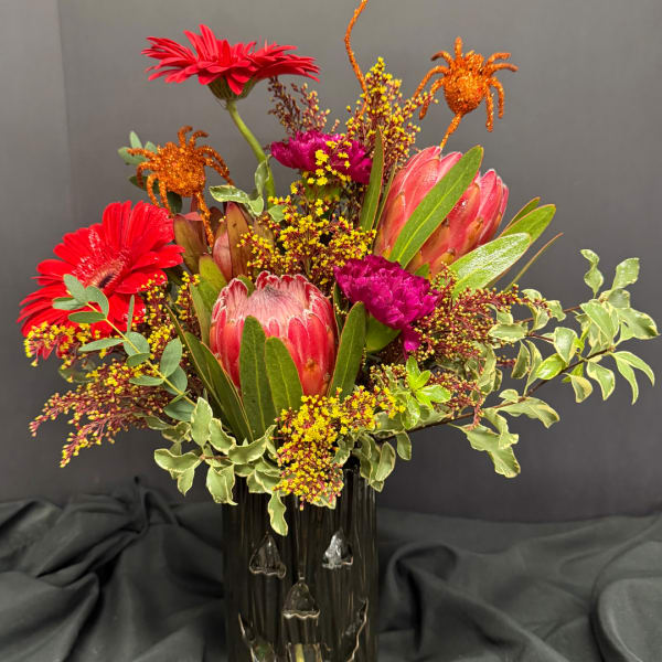 Bright mixed bouquet with red gerberas, pink protea, and orange spider accents in a dark vase