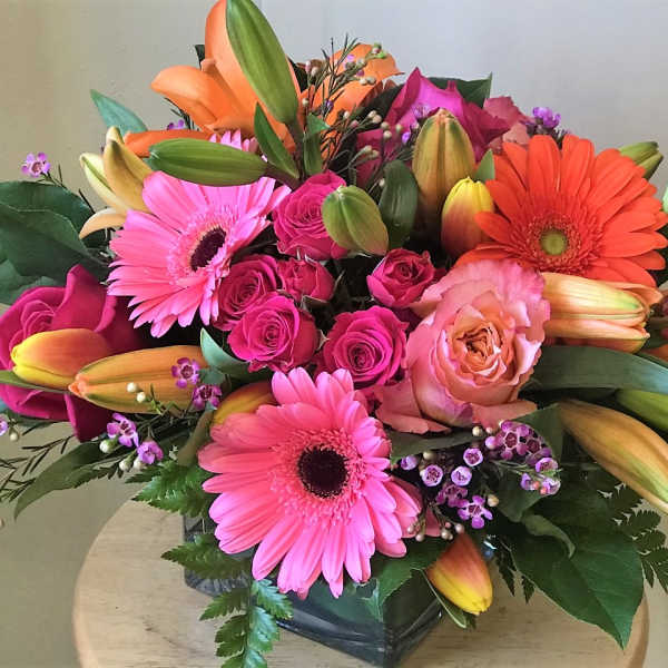 Bright mixed bouquet with pink gerberas, roses, and orange lilies in a glass vase.