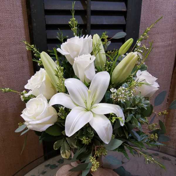 Arrangement of white lilies and roses in a clear glass vase with a burlap bow.