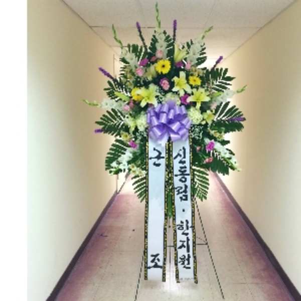Tall standing spray with yellow lilies and daisies, pink blooms, and a purple bow on an easel with Korean ribbons