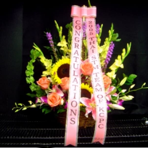 Basket arrangement with sunflowers, pink roses and lilies, accented by tall blooms and pink congratulations ribbons