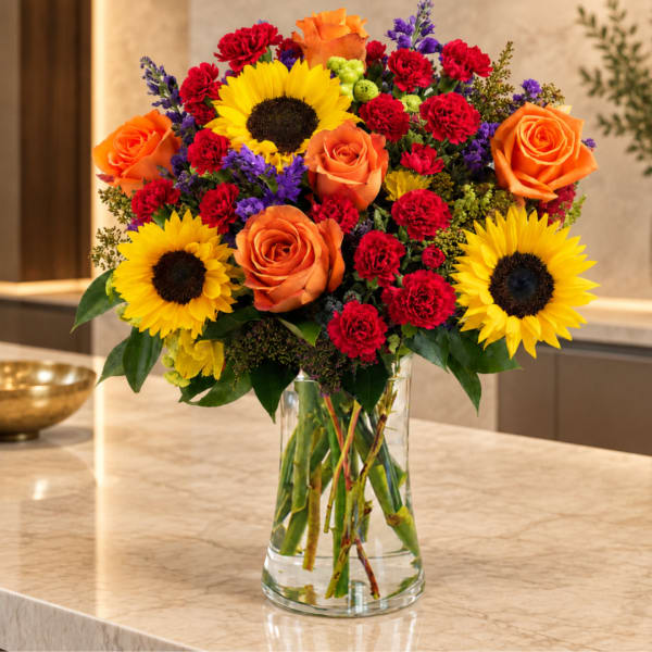 Bright mixed bouquet of sunflowers, orange roses, and red carnations in a glass vase