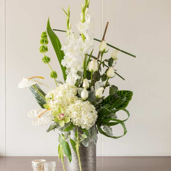 Tall white floral arrangement in a silver vase with candles beside it