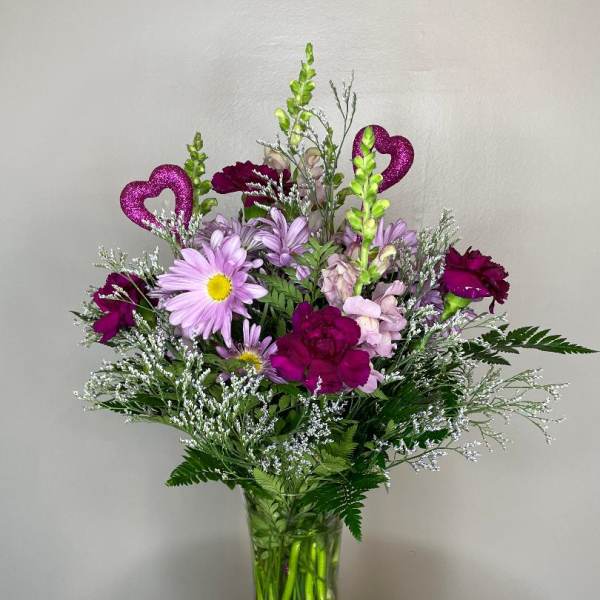 Tall vase arrangement of purple carnations, lavender daisies, and pink flowers with glitter heart accents.