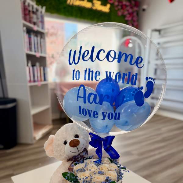 Cream teddy bear with box of white roses and clear balloon with blue text and balloons.
