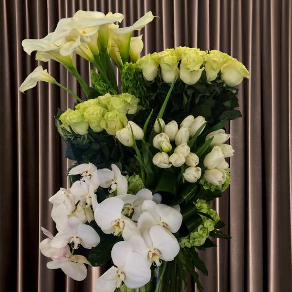 Tall white floral arrangement with calla lilies, roses, and orchids in a glass vase