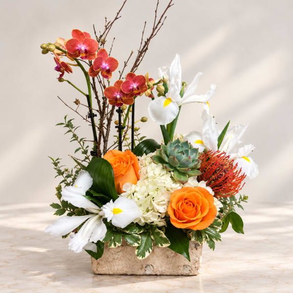 Mixed floral arrangement with orange roses, white irises, and orchids in a stone container
