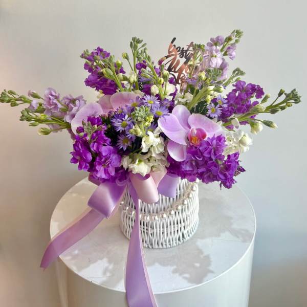 Purple and white floral arrangement in a white wicker basket with a lavender ribbon