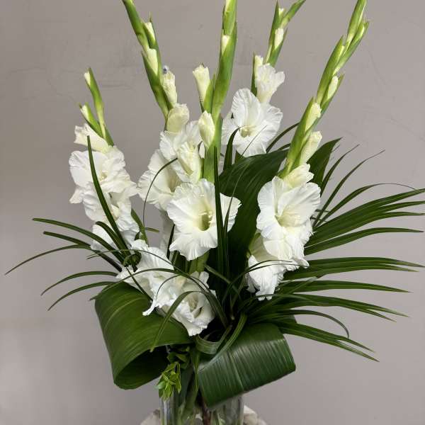 White gladiolus arrangement in a clear glass vase with broad green leaves