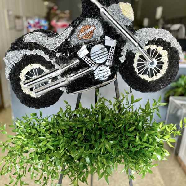 Motorcycle-shaped floral tribute with black, white, and gray flowers on an easel