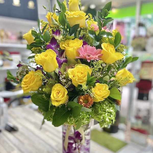 Tall arrangement of yellow roses, pink daisies, and orchids in a clear cylinder vase
