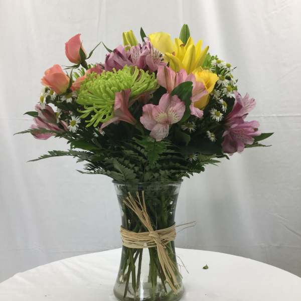Mixed bouquet of yellow lilies, peach roses, and pink blooms in a clear vase with a raffia tie