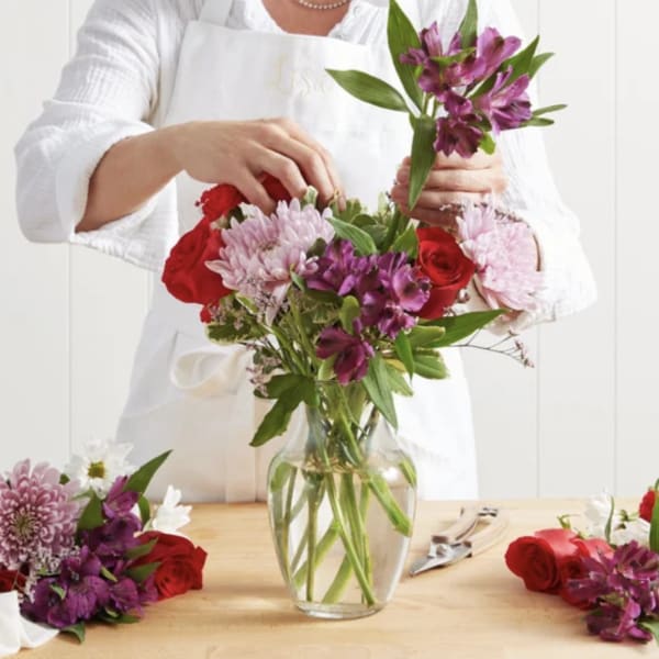 Person arranging a bouquet of red, pink, and purple flowers in a glass vase
