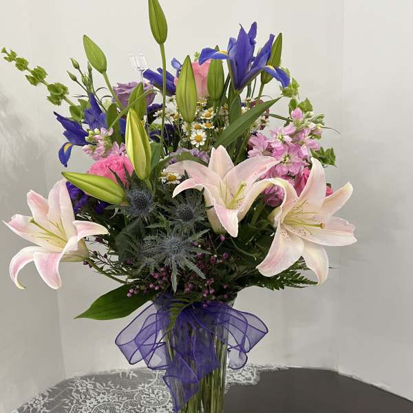 Mixed bouquet of lilies, irises, and pink flowers in a glass vase with a purple ribbon