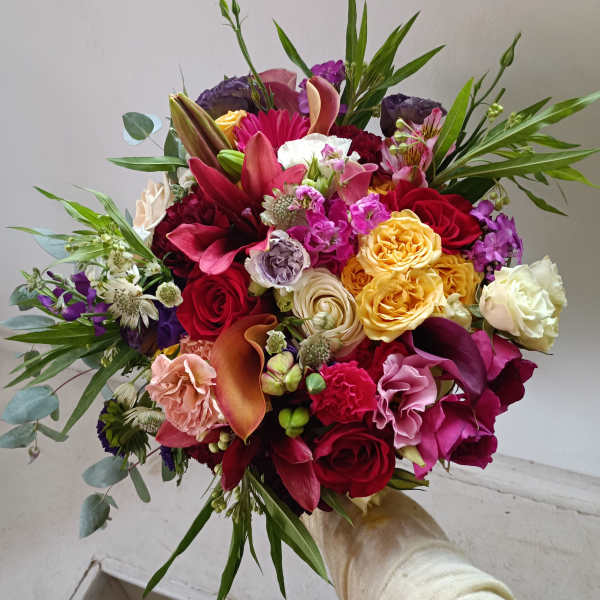 Handheld bouquet of mixed roses, lilies, and calla lilies in bright pink, red, yellow, and white.