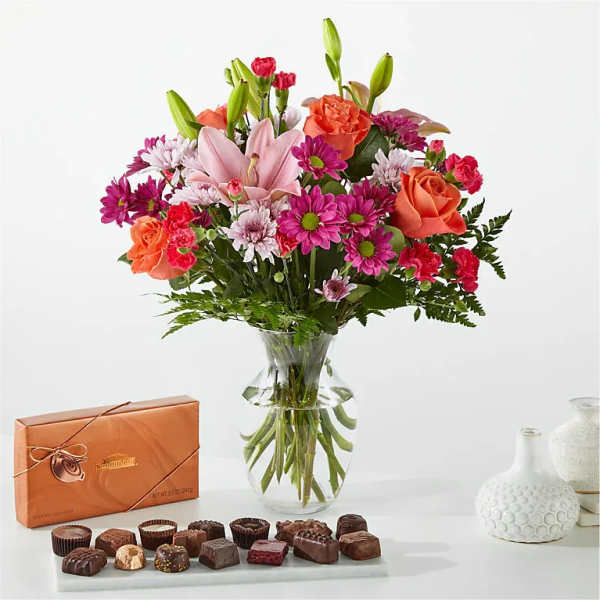Mixed bouquet of pink lilies, roses, and daisies in a glass vase with chocolates