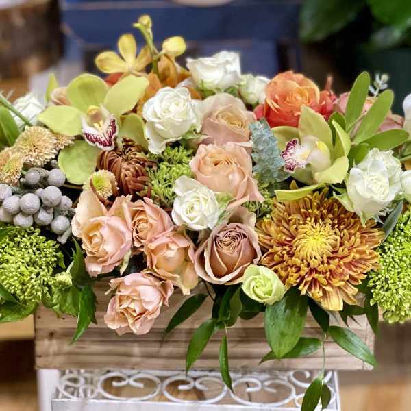 Low wooden box arrangement of peach roses, orchids, and mums in warm mixed colors
