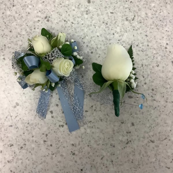 Two white rose boutonnieres with blue ribbon accents on a speckled floor