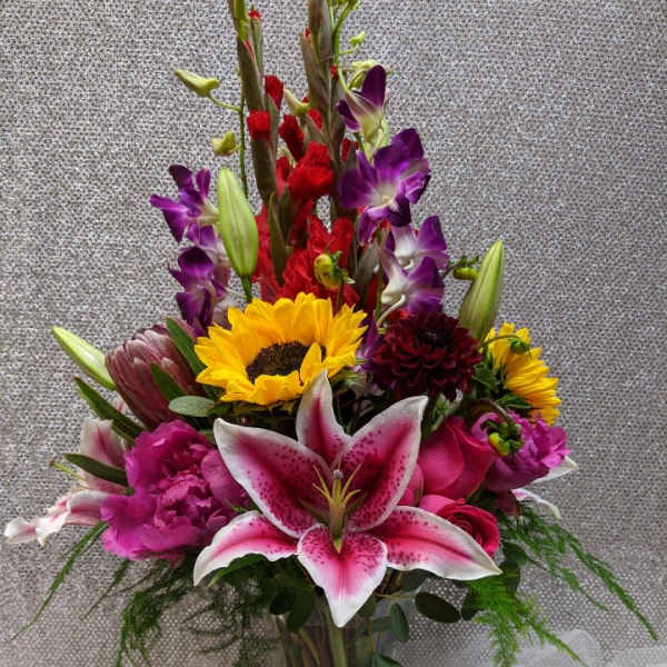 Tall mixed bouquet with pink lilies, sunflowers, purple orchids, and red blooms in a clear glass vase