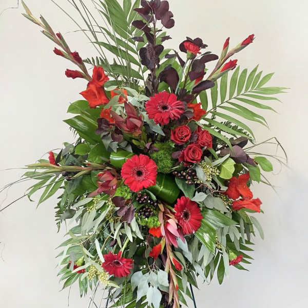 Tall standing spray of red gerbera daisies and roses with dramatic greenery on an easel.