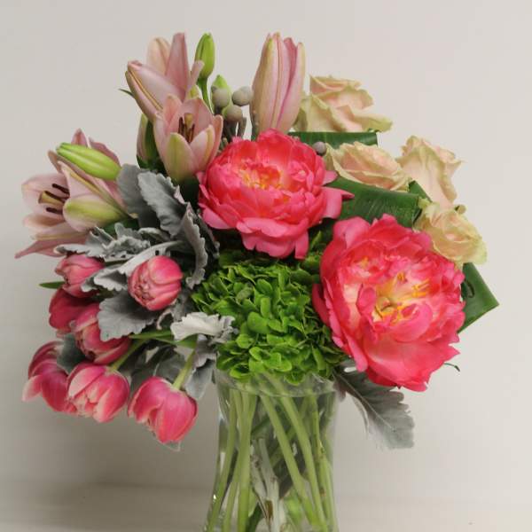 Pink peonies, tulips, lilies, and green hydrangea arranged in a clear glass vase