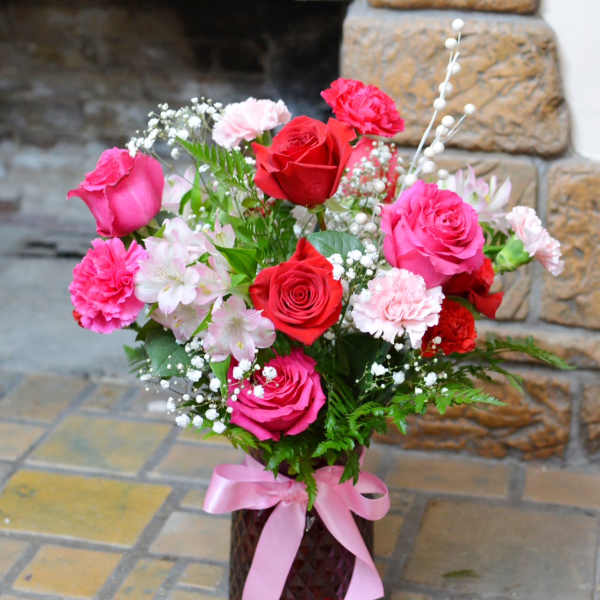 Pink and red roses with carnations in a vase tied with a pink ribbon.
