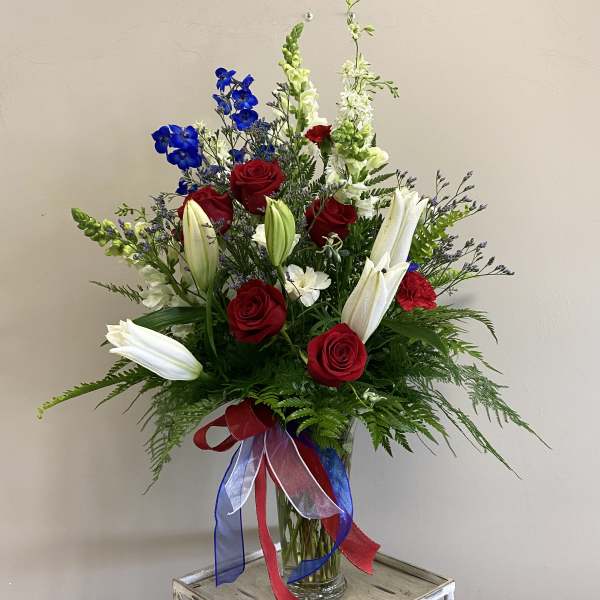Red roses and white lilies in a clear glass vase with red, white, and blue ribbons