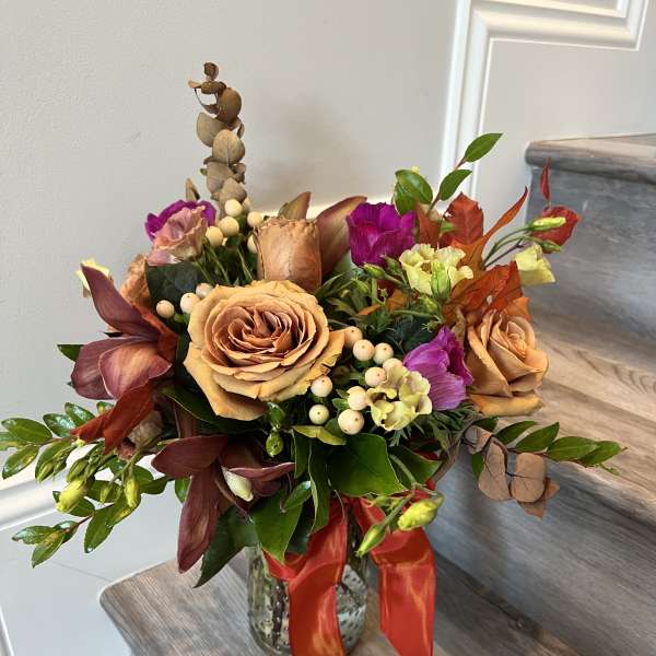 Mixed bouquet of peach roses and purple flowers in a glass vase with a red ribbon.