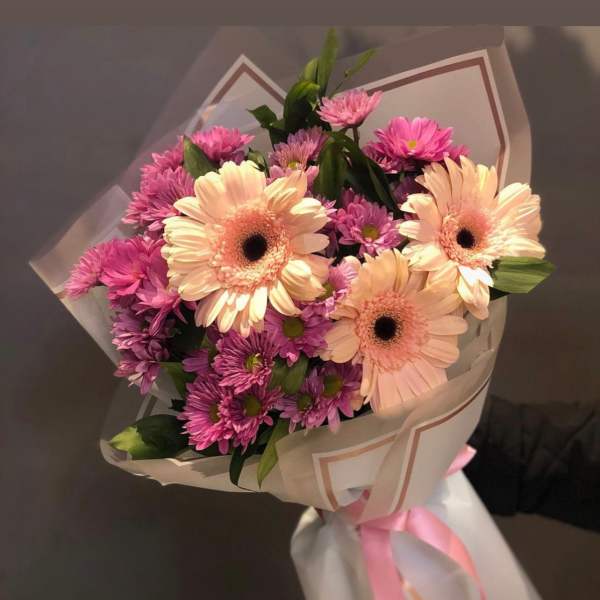Hand-tied bouquet of pale peach gerbera daisies and purple mums wrapped in white paper with a pink ribbon.