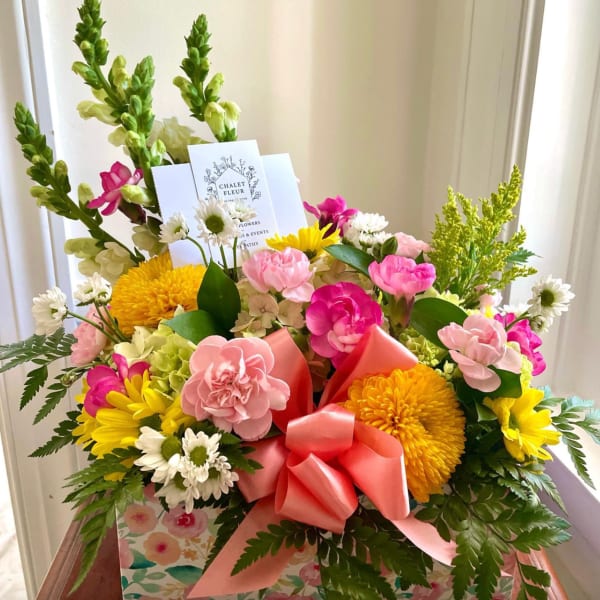 Colorful box arrangement of pink carnations, yellow mums, and white daisies with a pink bow and card.