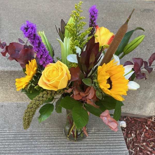 Mixed bouquet with yellow roses, sunflowers, and purple spikes in a glass vase