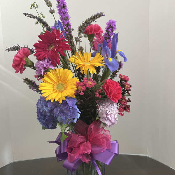 Tall mixed arrangement with yellow gerbera, blue hydrangeas, pink carnations and purple blooms in a glass vase.